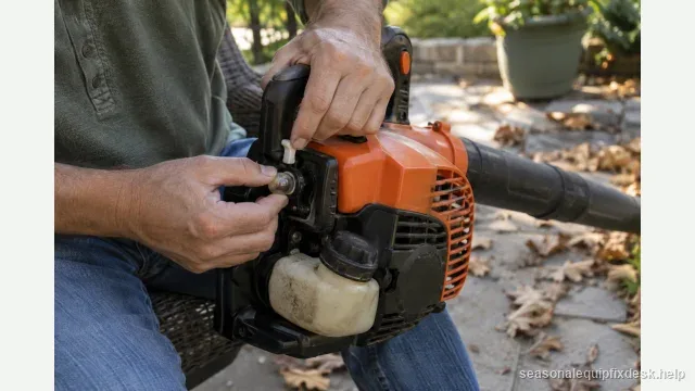A handheld gas leaf blower held across a person’s lap on a back patio, one thumb pressing the clear primer purge bulb on the carburetor while the other hand holds the choke lever at full choke, dry leaves on the ground.
