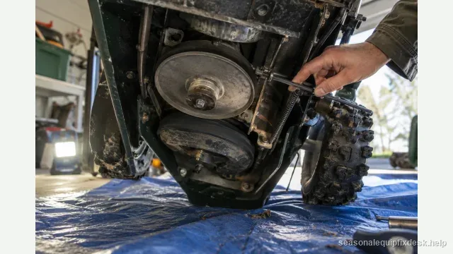 A snow blower tipped up onto its auger housing on a blue tarp in a garage, the underside exposed showing the rubber friction drive disc against the drive plate, a hand tracing the drive control cable.